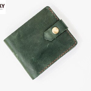 May include: A dark green leather wallet with a brown stitch detail and a gold button closure. The wallet has a flap closure with a strap.
