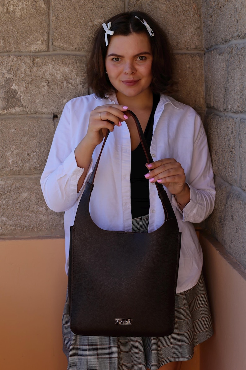 May include: A dark brown shoulder bag with a curved top and a long strap. The bag is held by a person wearing a white shirt and a black top. The bag has a small logo at the bottom.