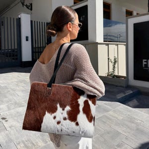 May include: A rectangular tote bag featuring a brown and white cowhide pattern with a textured fur finish. The bag has two thin black shoulder straps attached to the top edge, providing a structured silhouette for carrying personal items.
