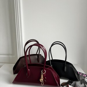 May include: Three stylish handbags in burgundy, black, and brown, each with a unique shape and double handles. The burgundy bag features a gold charm. Sunglasses and a magazine are on the white surface.