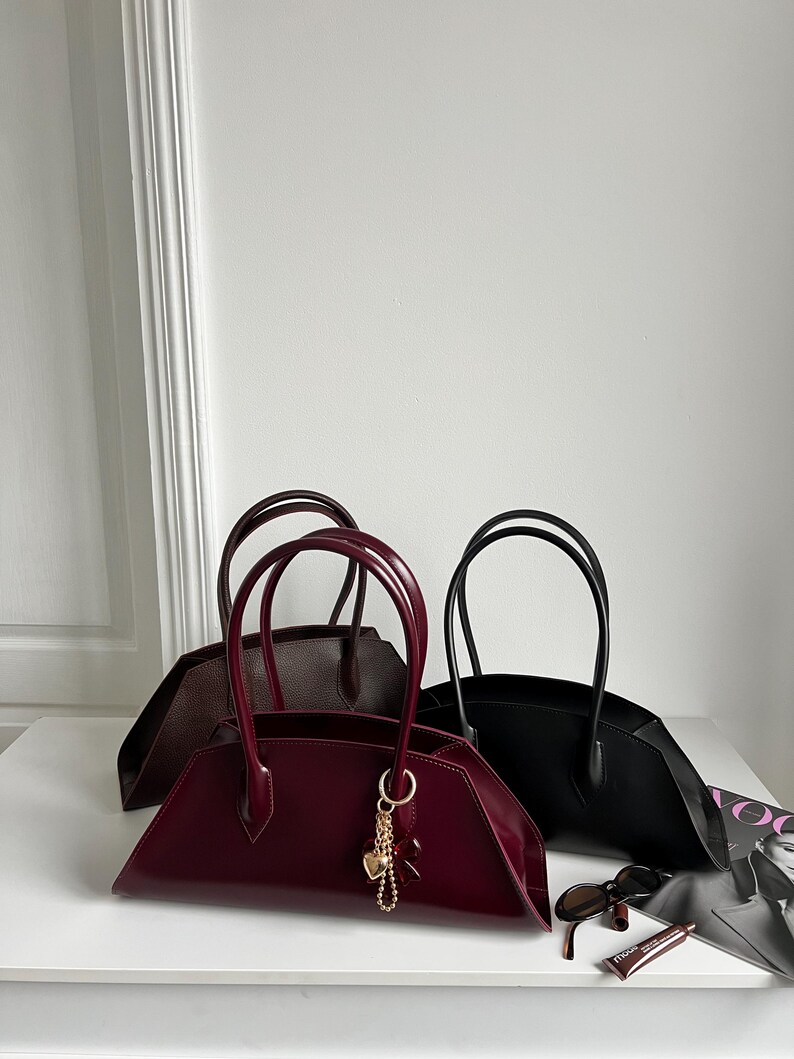 May include: Three stylish handbags in burgundy, black, and brown, each with a unique shape and double handles. The burgundy bag features a gold charm. Sunglasses and a magazine are on the white surface.