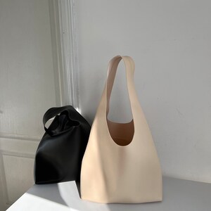May include: Two tote bags, one black and one cream, made of leather. The bags have a simple, minimalist design with a single handle.