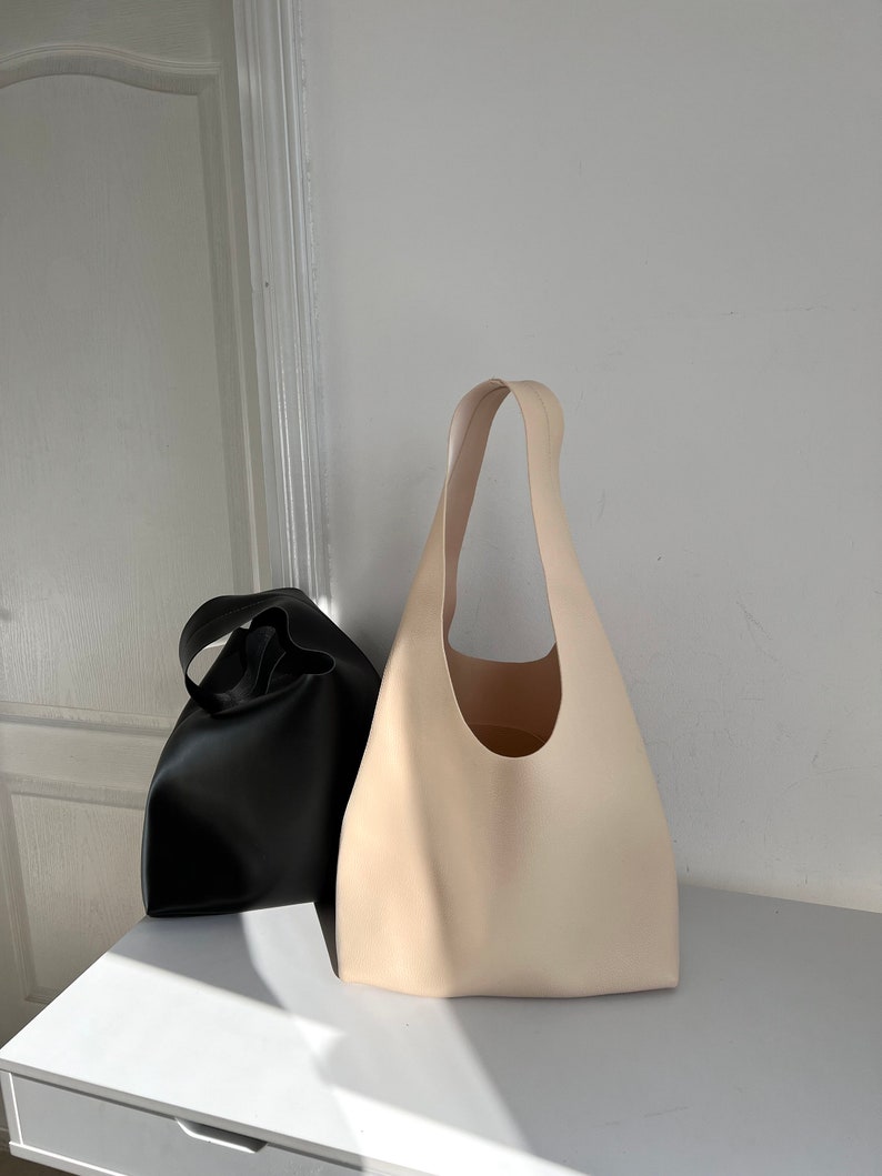 May include: Two tote bags, one black and one cream, made of leather. The bags have a simple, minimalist design with a single handle.