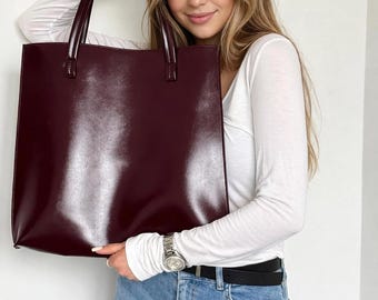 Könnte beinhalten: Eine weinrote Tragetasche mit glänzendem Finish und zwei Griffen. Die Tasche hat eine rechteckige Form. Die Tasche wird von einer Person gehalten, die ein weißes Langarmshirt und eine blaue Jeans trägt.