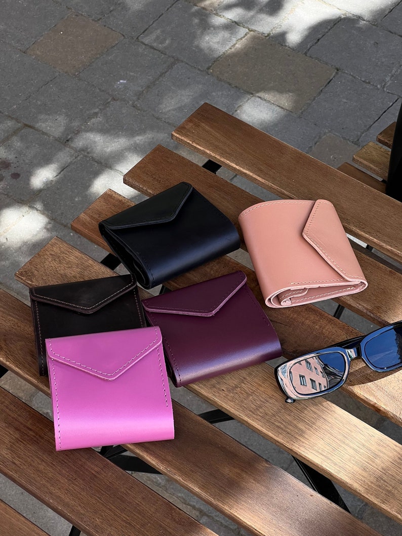 May include: Five leather wallets in different colors, including black, brown, pink, purple, and peach, are arranged on a wooden surface. A pair of black sunglasses is also on the surface.
