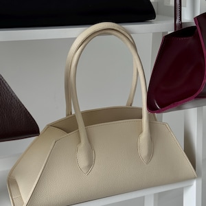 May include: A cream-colored handbag with a structured, geometric design. The bag features a curved top handle and a trapezoidal shape. Other handbags in black and burgundy are visible on the shelves.