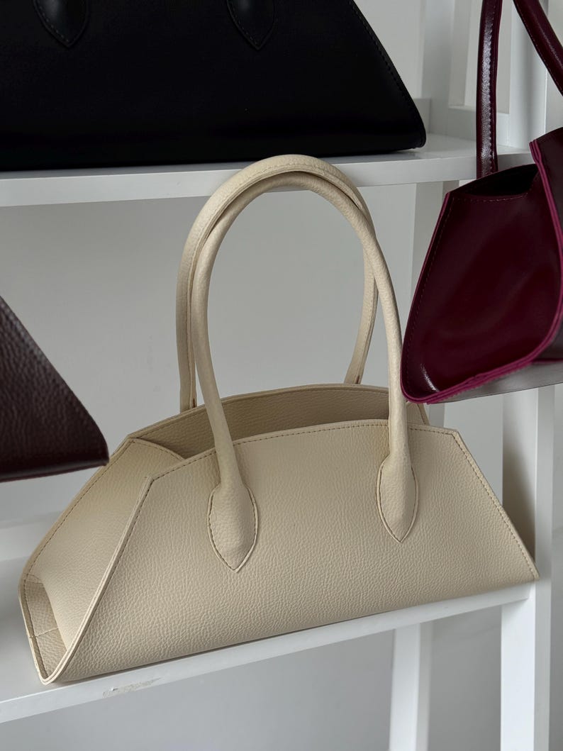 May include: A cream-colored handbag with a structured, geometric design. The bag features a curved top handle and a trapezoidal shape. Other handbags in black and burgundy are visible on the shelves.