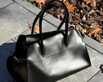 May include: A black leather handbag with two rounded handles. The bag has a rectangular shape with a slightly curved top and a zipper closure. The bag is sitting on a gray stone surface with fallen leaves in the background.
