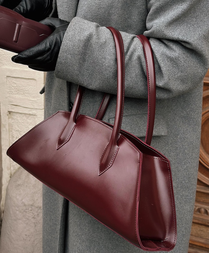 May include: A burgundy leather handbag with a structured design and long handles. The bag has a rectangular shape with a pointed top and is held by a person wearing black gloves and a gray coat.