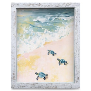 May include: A framed painting depicts a beach scene with three blue sea turtles on the sand. The artwork features a light blue ocean with white waves, a sandy beach, and a distressed white wooden frame.