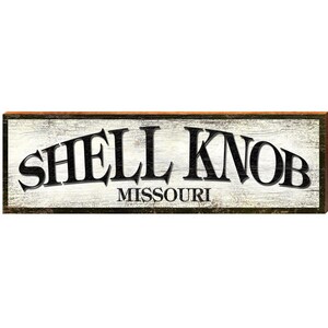 May include: A rectangular wooden sign with the words "SHELL KNOB" in large, black, arched letters above "MISSOURI" in smaller, black letters. The background is white with a wood grain texture, and the frame is brown.