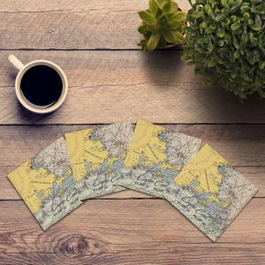 May include: Four square coasters with a vintage nautical chart design of Cape Cod, Massachusetts. The coasters are yellow and blue with the text "Cape Cod" and coordinates. A cup of coffee and a small plant are also visible.