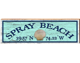 May include: A rectangular wooden sign with a distressed light blue finish and a dark blue border. The text reads SPRAY BEACH with the coordinates 39.57 N 74.22 W. A single tan seashell is centered between the coordinates.