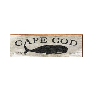 May include: A rectangular wooden sign with a weathered white finish featuring the black text CAPE COD in a serif font. Below the text is a black silhouette of a whale. The coordinates 41.66 N and 70.29 W are printed in black.