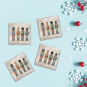 May include: Four square coasters with a distressed wood-look background feature colorful nutcracker soldier designs. Each coaster displays a different arrangement of the soldiers in red, green, blue, and black uniforms. The coasters are set against a light blue backdrop with snowflake and berry accents.
