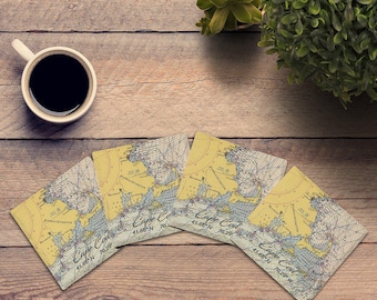 May include: A set of four square coasters featuring a nautical map of Cape Cod, Massachusetts. Each coaster displays the text Cape Cod with coordinates 41.66 N 70.29 W, set against a yellow and blue cartographic background with grid lines.