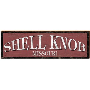 May include: A rectangular wooden sign with the words "SHELL KNOB" in large, white, arched letters above "MISSOURI" in smaller white letters. The background is a distressed red, with a black and brown border.