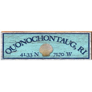 May include: Wooden sign with the text "QUONOCHONTAUG, RI" in dark blue, with a light blue background and a seashell in the center. Coordinates 41.33 N and 71.70 W are below the text. The sign has a dark blue border and a brown frame.