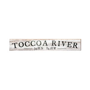 May include: A rectangular wooden sign with the words "Toccoa River" in black, distressed lettering. Below the text are the coordinates 34.88 N 84.28 W. The sign has a white background with a weathered appearance.