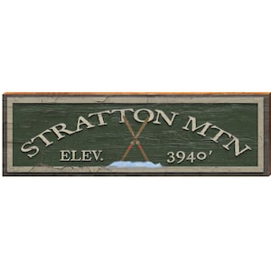 May include: Wooden sign with the words "STRATTON MTN" in white, arched across the top. Below, "ELEV. 3940'" is displayed. The sign features crossed skis and a snow scene, with a green background and a distressed wood frame.