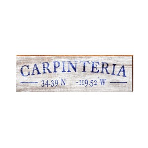 May include: A rectangular wooden sign with the word "CARPINTERIA" in blue, distressed lettering. Below the word are the coordinates 34.39 N -119.52 W. The sign has a weathered, rustic appearance, with a light brown border.