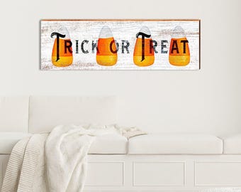 Könnte beinhalten: Ein rechteckiges Holzschild mit weißem Used-Look-Finish und dem Schriftzug TRICK OR TREAT in schwarzen Großbuchstaben. Vier Illustrationen von gelb-orange-weißen Zuckermais-Bonbons sind hinter den Buchstaben platziert.