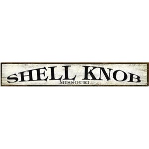 May include: A rectangular wooden sign with the words "SHELL KNOB MISSOURI" in black, distressed lettering. The sign has a white background with a weathered appearance and a dark brown border.