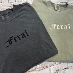 Puede incluir: Dos camisetas con la palabra "Feral" bordada. La camiseta de la izquierda es gris oscuro con bordado negro. La camiseta de la derecha es verde claro con bordado blanco.