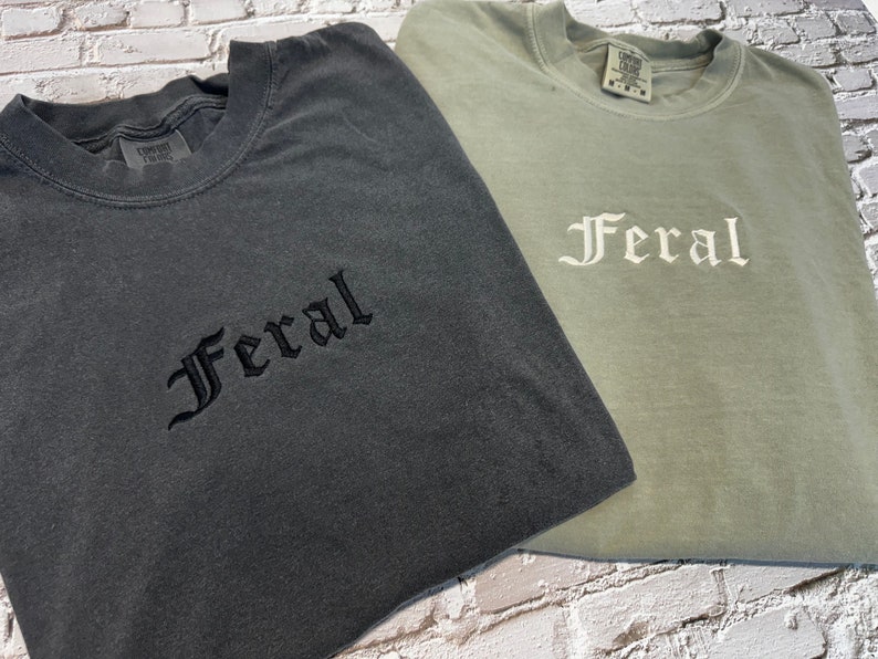 Puede incluir: Dos camisetas con la palabra "Feral" bordada. La camiseta de la izquierda es gris oscuro con bordado negro. La camiseta de la derecha es verde claro con bordado blanco.