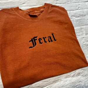Puede incluir: Una camiseta marr&oacute;n con la palabra "Feral" bordada en negro en una fuente g&oacute;tica.