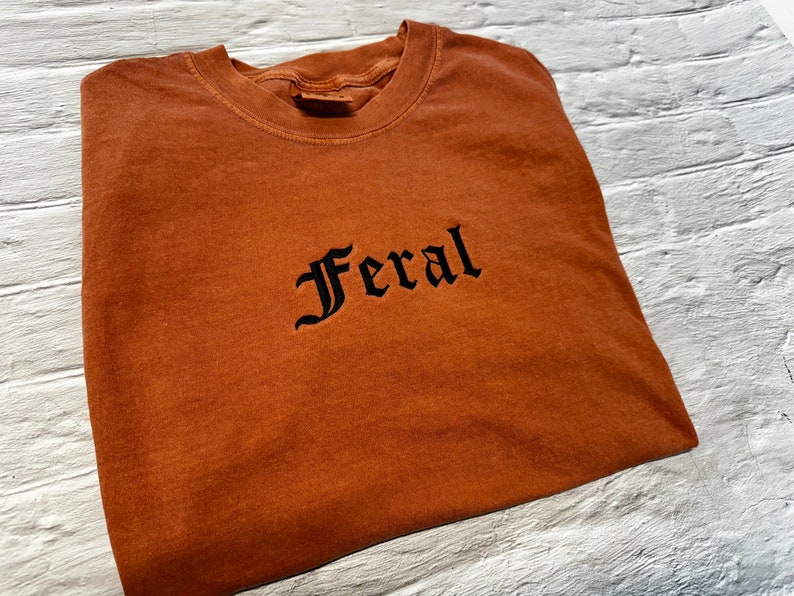 Puede incluir: Una camiseta marr&oacute;n con la palabra "Feral" bordada en negro en una fuente g&oacute;tica.
