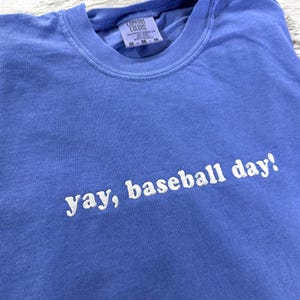 May include: A light blue t-shirt with the text "yay, baseball day!" embroidered in white.