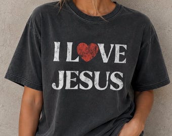 May include: A charcoal grey short-sleeve t-shirt featuring the white distressed text I LOVE JESUS with a red heart replacing the word LOVE. The shirt has a crew neckline and a relaxed fit with a faded, vintage-inspired wash throughout the fabric.