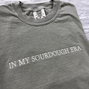 May include: A gray t-shirt with the text "IN MY SOURDOUGH ERA" embroidered in white on the front. The shirt is made of a soft, comfortable material and has a relaxed fit.