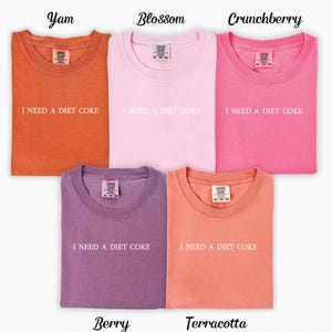 Op de afbeelding: Vijf T-shirts in verschillende kleuren met de tekst "Ik heb een light cola nodig" gedrukt op de voorkant. De kleuren zijn: Yam, Blossom, Crunchberry, Berry en Terracotta.