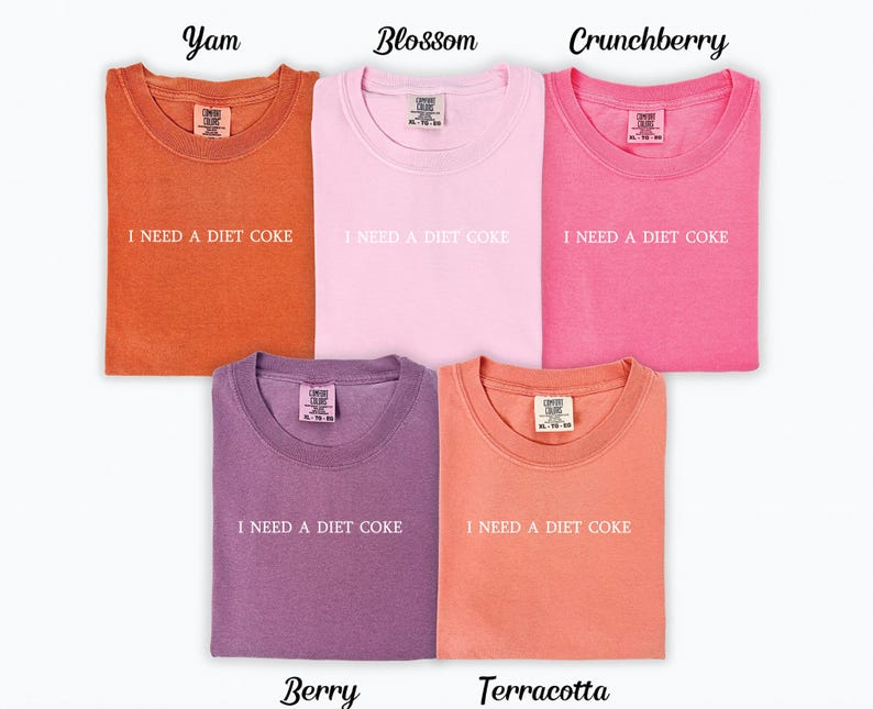 Op de afbeelding: Vijf T-shirts in verschillende kleuren met de tekst "Ik heb een light cola nodig" gedrukt op de voorkant. De kleuren zijn: Yam, Blossom, Crunchberry, Berry en Terracotta.
