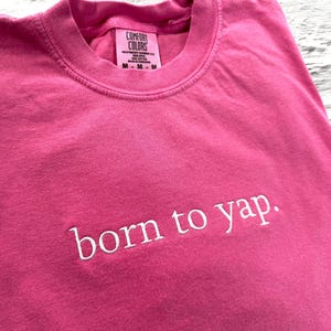 May include: Pink t-shirt with the text "born to yap." in white embroidery.
