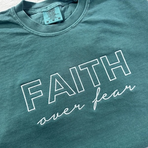 Puede incluir: Camiseta de color verde azulado con las palabras "FAITH over fear" bordadas en blanco. Cuello redondo y etiqueta "Comfort Colors" visibles. La tela parece suave y ligeramente descolorida. Una prenda casual.