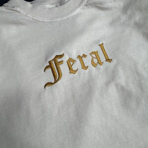 Puede incluir: Camiseta blanca de manga larga con la palabra "Feral" bordada en oro en una fuente g&oacute;tica.