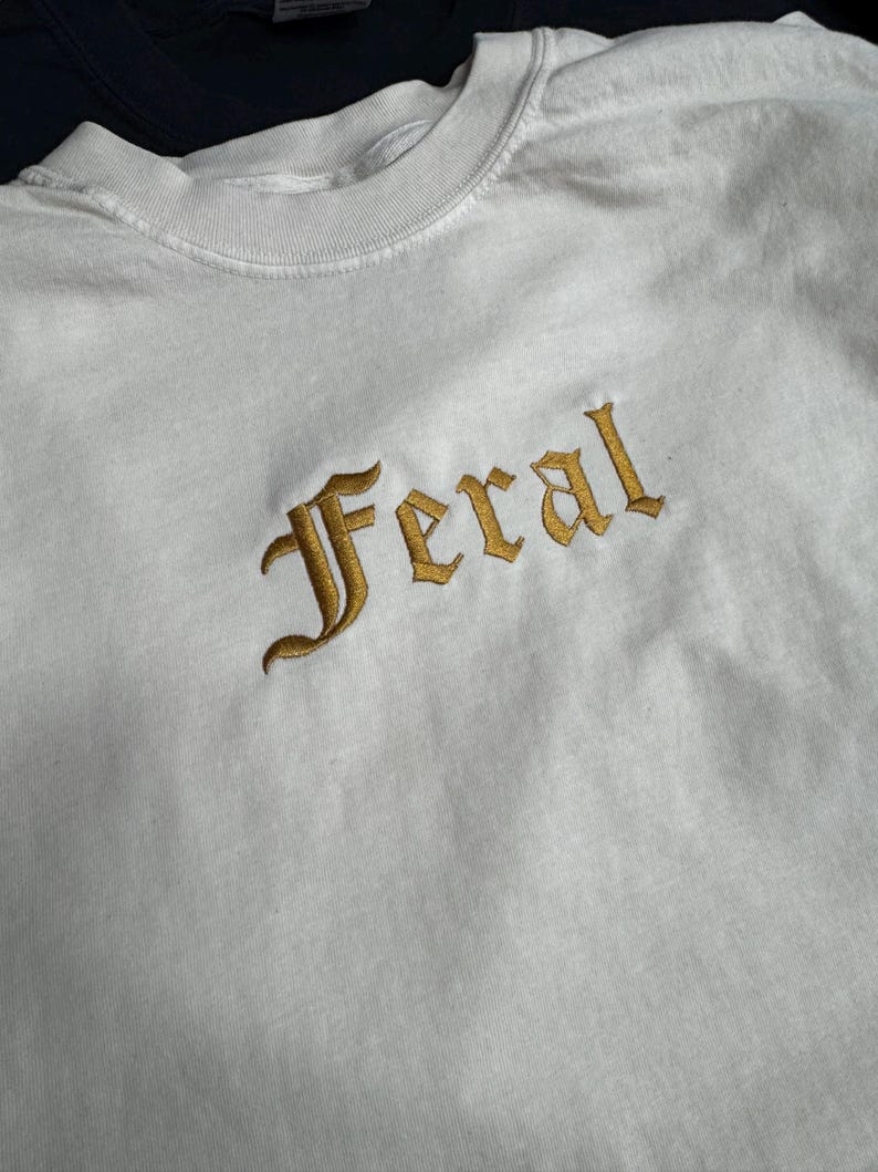 Puede incluir: Camiseta blanca de manga larga con la palabra "Feral" bordada en oro en una fuente g&oacute;tica.