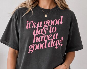 May include: A charcoal grey short-sleeved t-shirt featuring the pink text "it's a good day to have a good day!" printed on the front. The shirt has a crew neckline and a relaxed fit, with a slightly faded, washed-out appearance to the fabric.