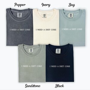 Op de afbeelding: Zes T-shirts in verschillende kleuren, waaronder peper, ivoor, baai, zandsteen en zwart. Elk shirt heeft de tekst "I NEED A DIET COKE" op de voorkant.