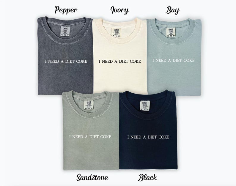 Op de afbeelding: Zes T-shirts in verschillende kleuren, waaronder peper, ivoor, baai, zandsteen en zwart. Elk shirt heeft de tekst "I NEED A DIET COKE" op de voorkant.