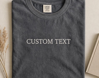 May include: A charcoal grey crew neck t-shirt with a vintage-washed finish. The shirt features the white embroidered text "CUSTOM TEXT" centred on the chest. A white brand label is sewn inside the collar with the text "COMFORT COLORS".