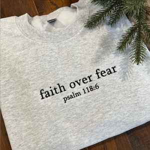 Op de afbeelding: Grijs sweatshirt met de tekst "faith over fear psalm 118:6" in zwart gedrukt.