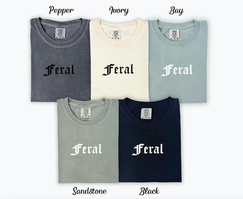 Puede incluir: Cinco camisetas de diferentes colores con la palabra "Feral" impresa en una fuente g&oacute;tica. Los colores son Pepper, Ivory, Bay, Sandstone y Black.