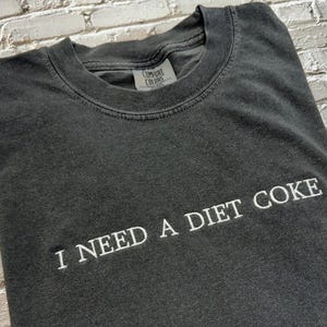 Op de afbeelding: Een donkergrijs T-shirt met de tekst "I NEED A DIET COKE" in wit.