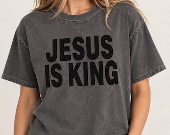 May include: A charcoal grey short-sleeved t-shirt with a washed, vintage-style finish. The front of the shirt features the bold, black, sans-serif text "JESUS IS KING" printed in two lines across the centre of the chest area.