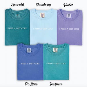 Op de afbeelding: Vijf T-shirts in verschillende kleuren met de tekst "I NEED A DIET COKE" erop gedrukt. De kleuren zijn smaragdgroen, chambray, violet, flo blauw en zeegroen.