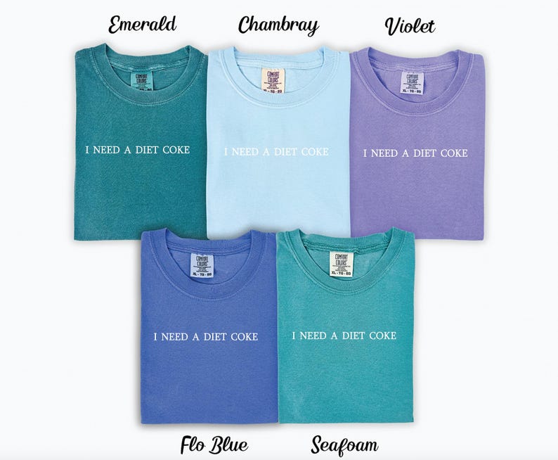 Op de afbeelding: Vijf T-shirts in verschillende kleuren met de tekst "I NEED A DIET COKE" erop gedrukt. De kleuren zijn smaragdgroen, chambray, violet, flo blauw en zeegroen.